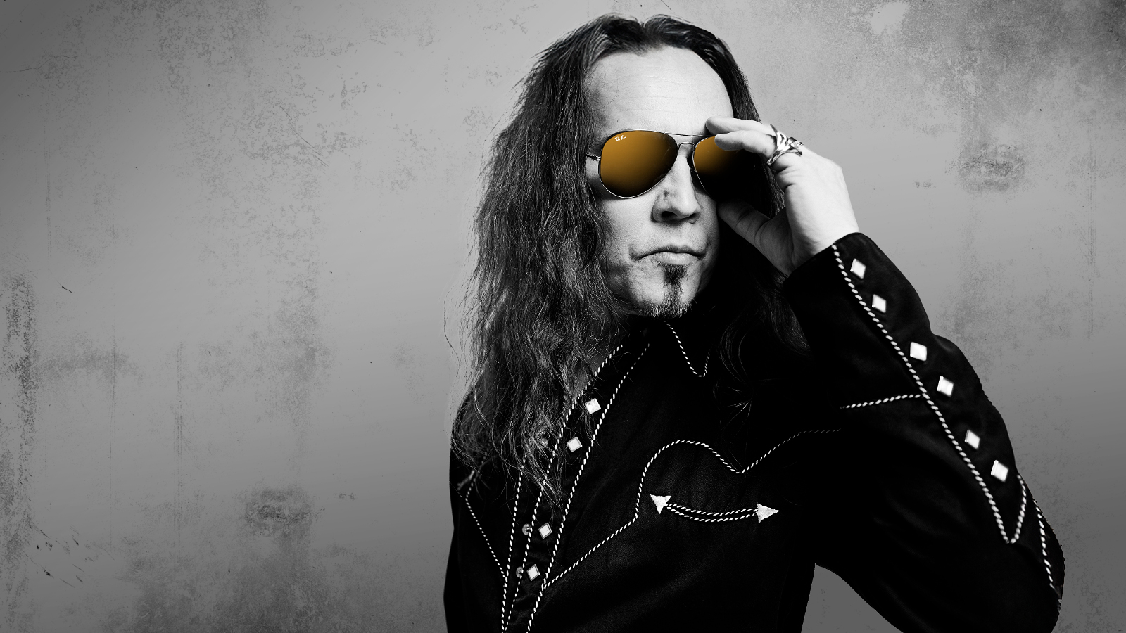 Kuvassa on Jukka ahola puolivartalokuvassa ja hänellä on musta takki päällään. Siinä on nappeja hihassa ja Ahola pitää vasemmalla kädellään aurinkolaseista kiinni. Kuva on musta-valkoinen tausta on harmaa.