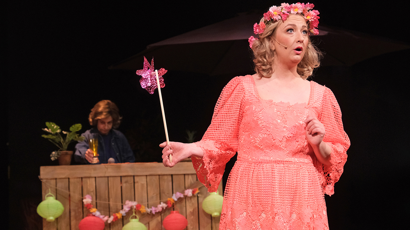 Kuvassa on keskellä nainen, jolla on oranssin punainen mekko päällään ja oikeassa kädessään violetti vappuhyrrä. Taustalla on puinen baaritiski ja sen takana mies, jolla on olutlasi kädessään.