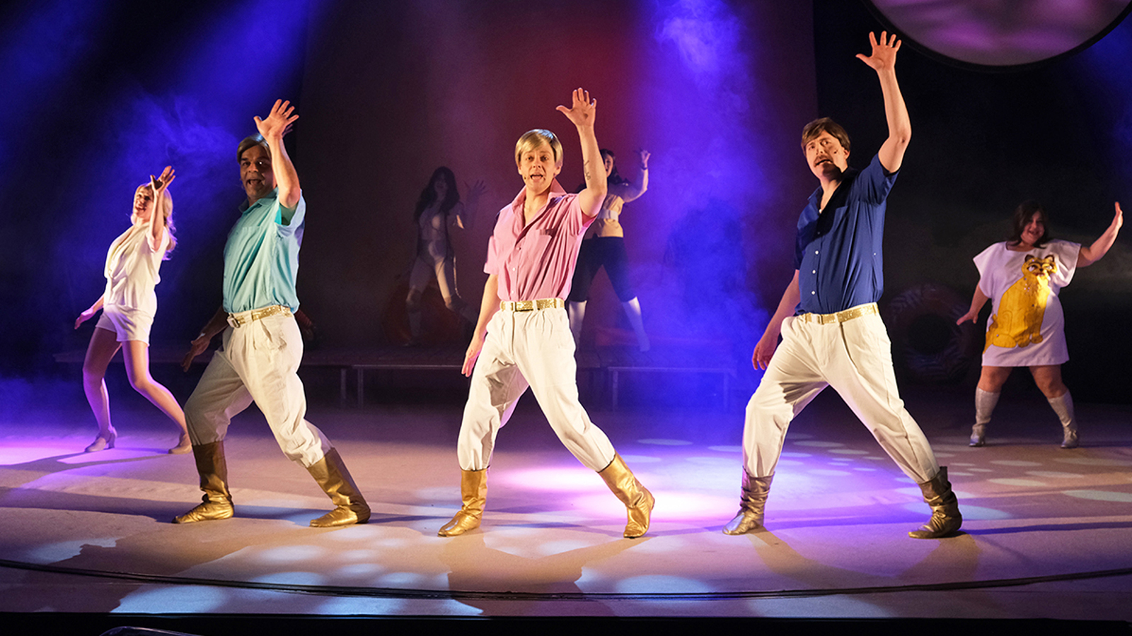 Kuvassa on keskellä kolme miestä tanssimassa vaaleissa housuissa, kultaisissa saappaissa ja eri värisissä paidoissa. Taustalla on himmeissä valoissa naistanssijoista. 