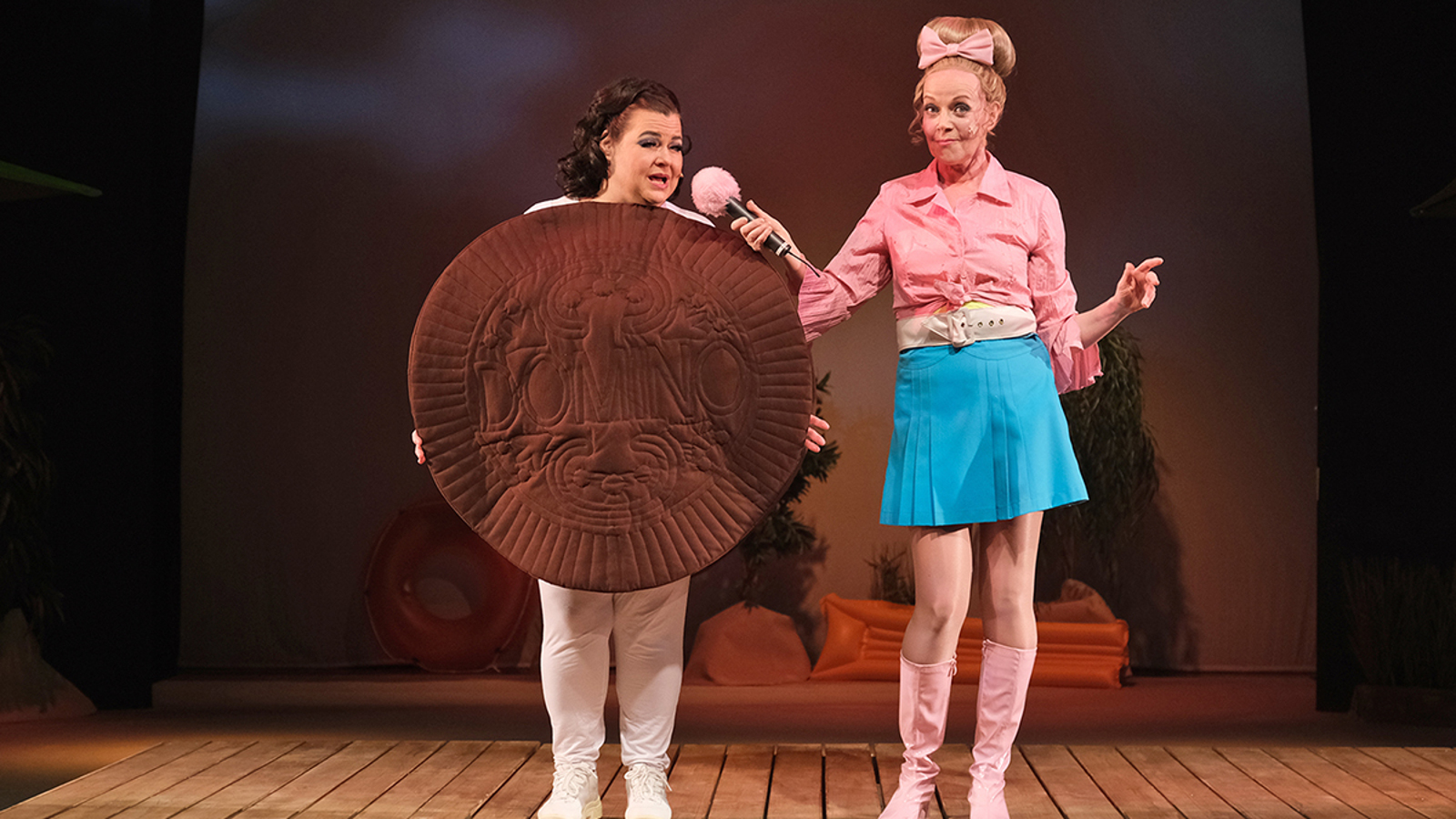 Kuvassa on kaksi naista seisomassa puuterassilla. Vasemmalla on nainen, joka pitelee isoa ruskeata keksin näköistä pöyrylää. Vieressä oikealla seisoo nainen vaalean punaisessa jakussa ja turkoosissa puolihameessa. Hän pitelee vaalean punaista mikrofonia toisen naisen edessä.