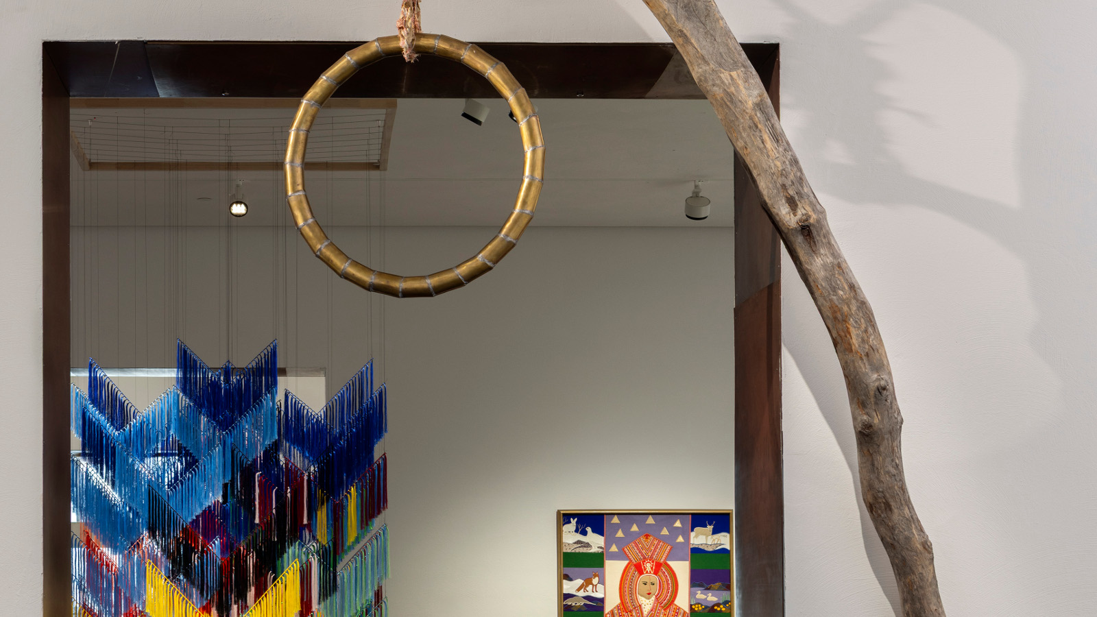 Kuvassa on etualalla kapeaa puunrunkoa ja siitä roikkuu puinen, pyöreä hirttosilmukka. Taustalla näkyy roikkuva tekstiilityö, jossa on Lapin värejä ja näyttelysalia.