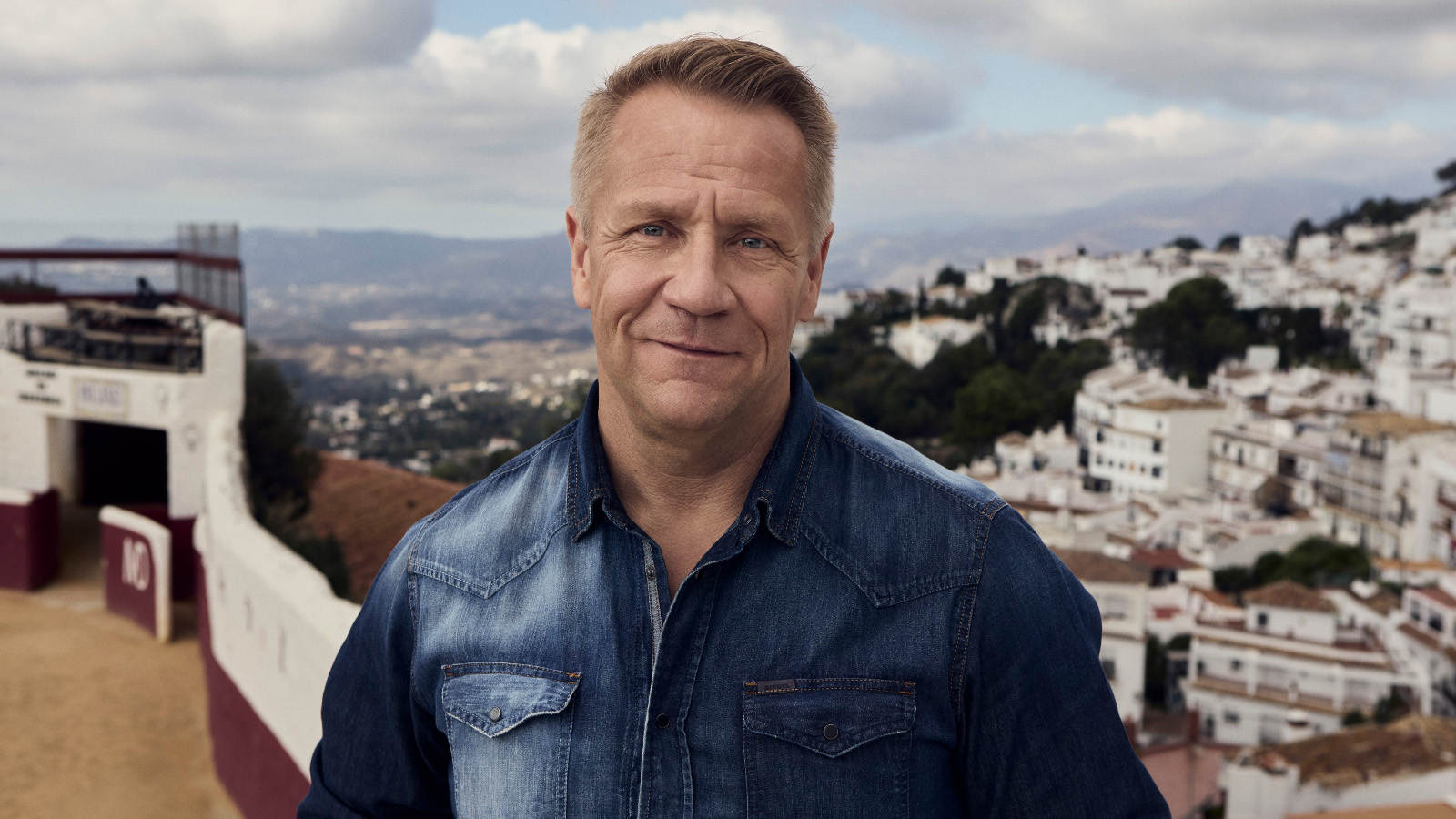 Kuvassa on Olli Lindholm seisomassa puolivartalokuvassa ja hänellä on tumman sininen farkkutakki päällään. Taustalla on valkoista kylää vuoren rinteellä ja puolipilvistä taivasta.