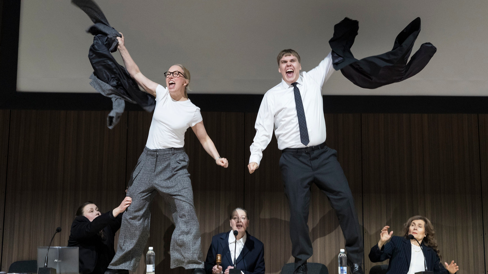 Kuvassa on nuori nainen vasemmalla ja hän heiluttaa takkiaan ja hänellä on valkoinen t-paita ja harmaat, löysät housut päällää, Oikealla on nuori mies, jolla on valkoinen paita jamusta kravatti sekä mustat housut päällään. Taustalla alhaalla on kolme henkilöä pöydän ääressä mustissa takeissa.