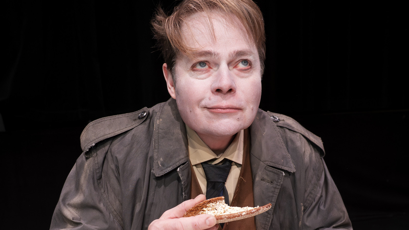 Kuvassa on mies ruskea päällystakki, kauluspaita ja kravatti päällään ja piirakka kädessään. Hän istuu.