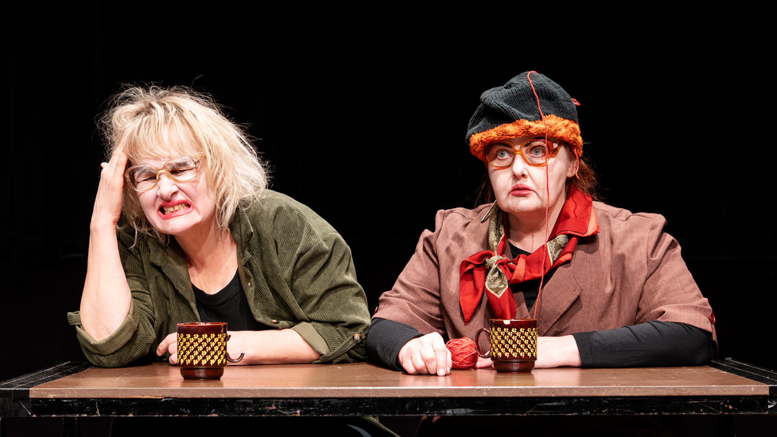 Kuvassa on kaksi naista istumassa pöydän ääressä ja heillä on kahvimukit edessään. Vasemmalla on vaaleatukkainen nainen, joka pitelee oikealla kädellään päätään. Oikealla on nainen mustassa baskerissa ja ruskeassa takissa ja hän tuijottaa eteenpäin.