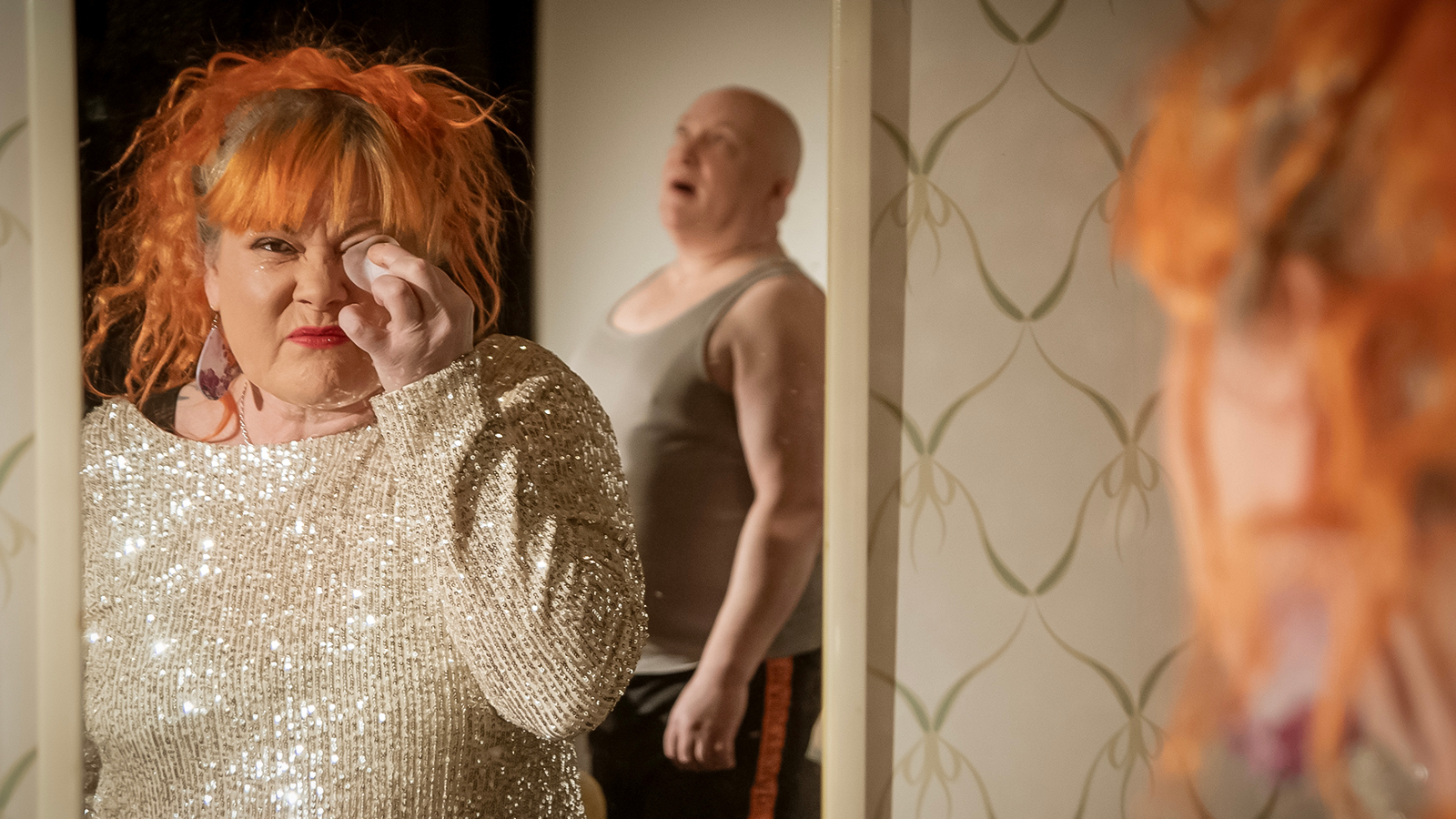 Kuvassa on Mira Lukkari oikealla osaksi selin ja hän näkyy vasemmalla peilistä poistamassa silmämeikkiä pienellä tyynyllä. Hänellä on vaalea glitterpusero päällää ja punaiset hiukset. Taustalla näkyy Jari Lukkari seisomassa sivuttain harmaassa, hihattomassa t-paidassa.