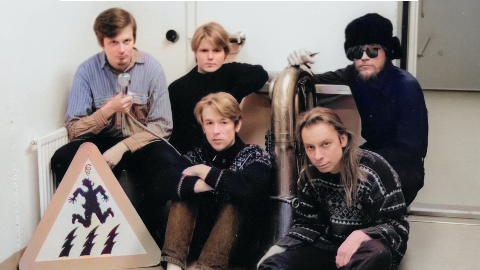 Kuvassa on viisi nuorta miestä, joista kolme istuu takarivissä ja heistä yhdellä on vaalean sininen pusero ja muilla mustat puserot. Edessä istuu kaksi miestä ja vasemmalla heidän vieressään on kolmioliikkennemerkki jossa on pomppivan henkilön kuva. Kuva on osa kirjan kannesta.