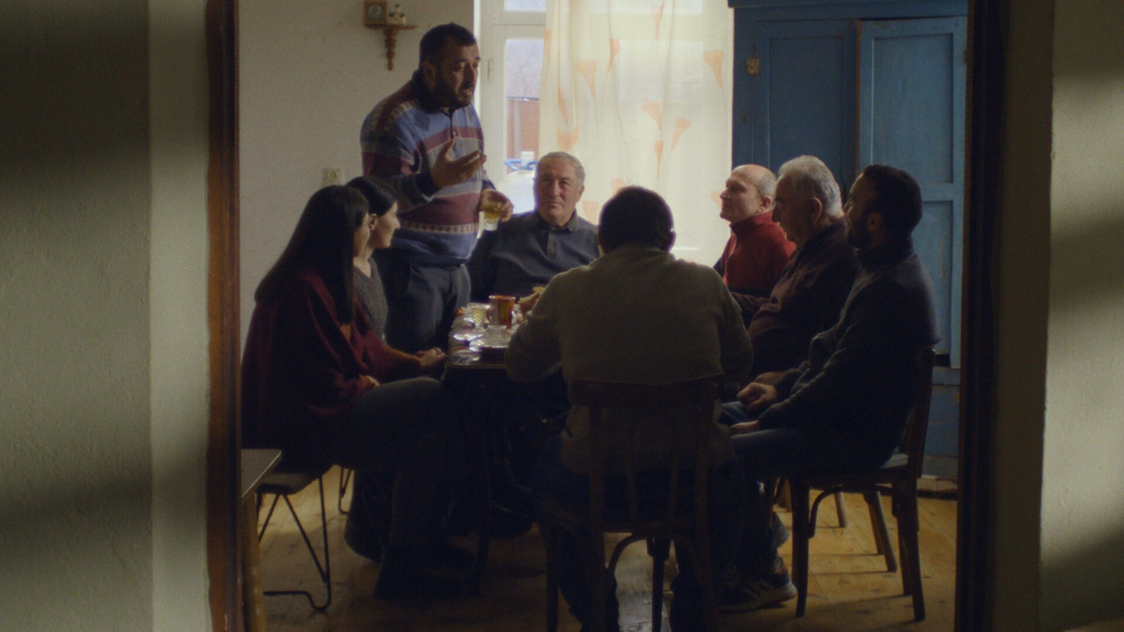 Kuvassa on pienessä huoneessa kahvipöydässä kahdeksan henkilöä. Pöydän päässä on vanhahko mies ja yksi mies seisoo hänen vieressään vasemmalla. Kuvan sävy on hämärä. 