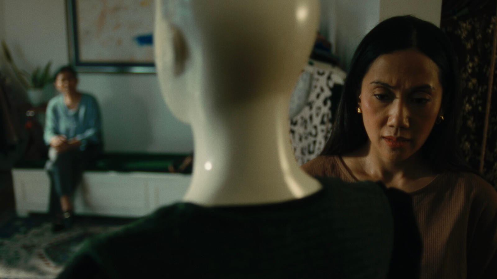  Kuvassa on etualalla mallinuken pää ja kaula takaapäin. Oikealla on naisen kasvot ja häne sovittaa jotakin nuken päälle. Taustalla istuu nainen ja hän näkyy epätarkasti. Kuvan sävy on vihertävä.