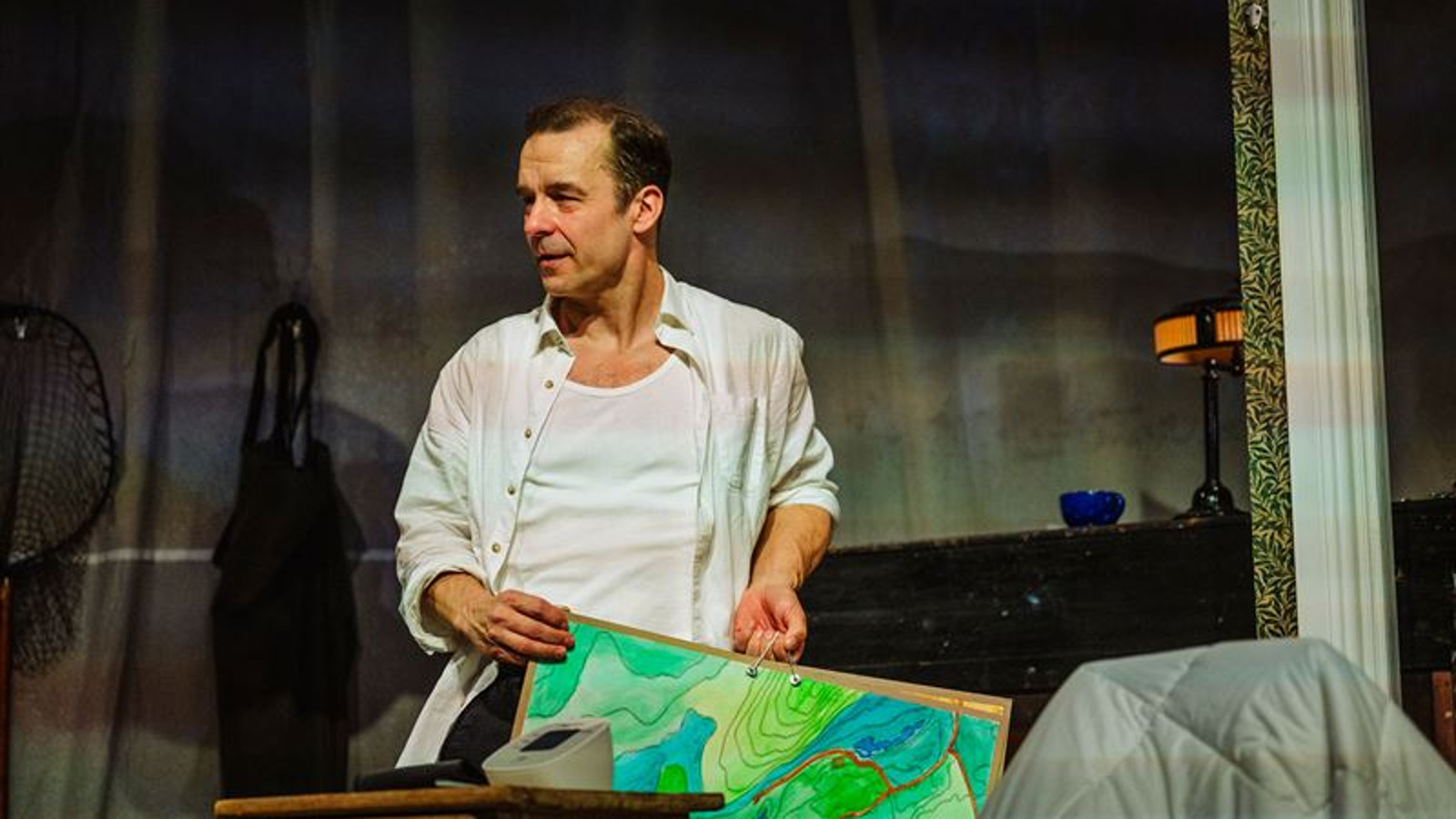 Kuvassa on Ilja Peltonen valkoisessa paidassa puolivartalokuvassa ja hänellä on käsissään vihreä maastokartta. Tausta on tumma.     