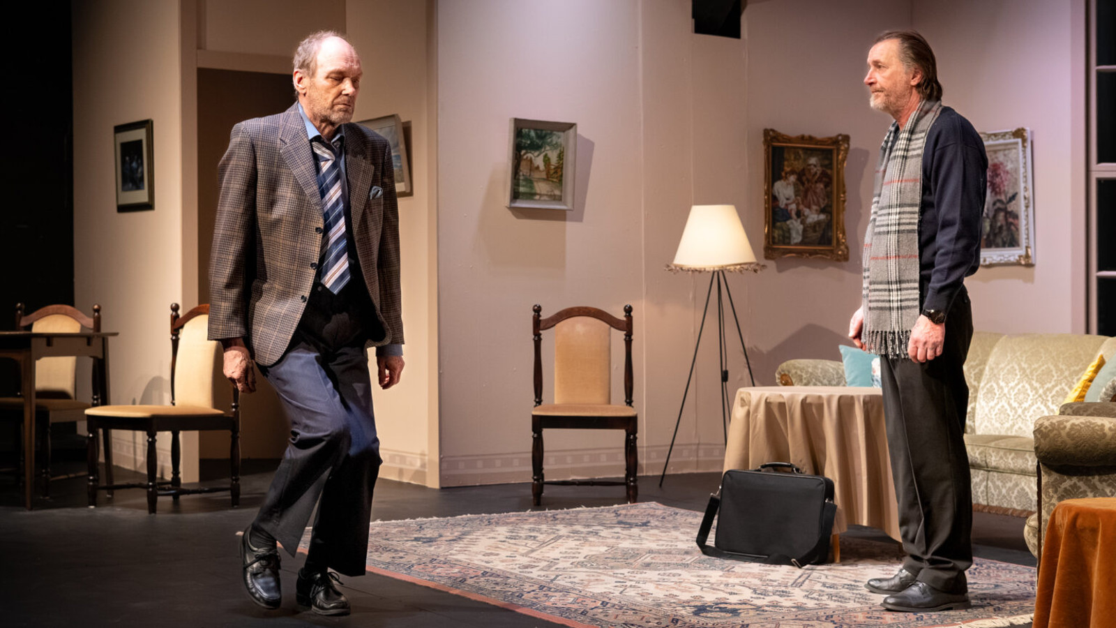 Vasemmalla on mies ruskeassa takissa ja sinisissä housuissa kävelemässä olohuoneessa. Oikealla seisoo sivuttain mies, joka katsoo kävelevää miestä.