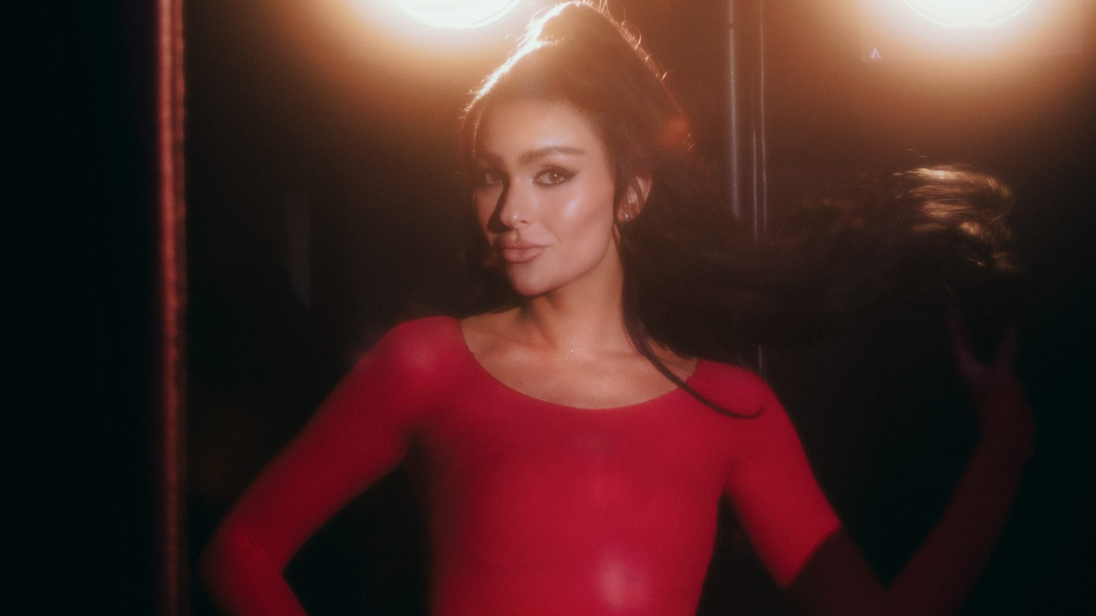 Kuvassa on keskellä Sara Siipola puolivartalokuvassa. Hänellä on punainen mekko päällään ja hänellä on tummat hiukset nutturalla. Taustalla tumman ruskeaa ja kaksi ylävaloa.