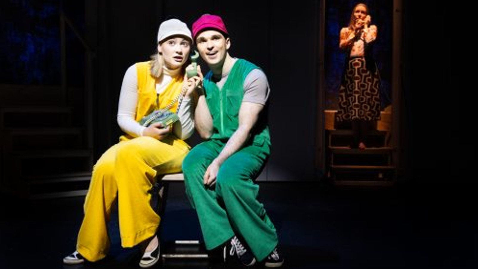 Kuvassa ovat istumassa penkillä vasemmalla nuori nainen keltaisessa haalarissa valkeassa lippiksessä ja oikealla nuori mies vihreässä haalarissa ja punaisessa lippiksessä. Tausta on tumma. Takana näkyy oikealla pienenä nainen, joka istuu tuolilla.