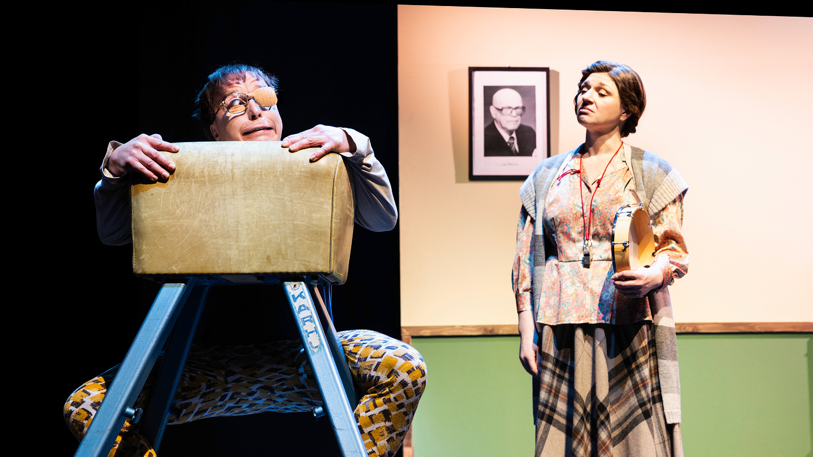 Kuvassa on mies ruskean hyppypenkin takana, josta hän pitää kiinni. Oikealla seisoo nainen ruskearuudullisessa mekossa. Taustalla on vaalean ruskeaa seinää, jossa on Urho Kekkosen muotokuva. 