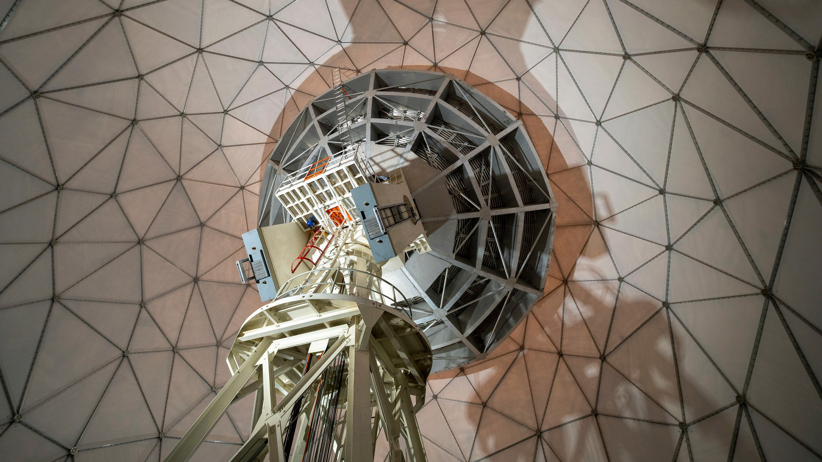 Kuvassa on avaruusteleskooppi kuvattuna alhaalta ylöspäin ja taustallaon rautaverkkoa vaaleanruskealla pohjalla.