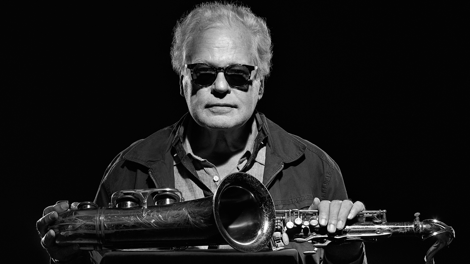 Kuvassa on Eero Koivistoinen puolivartalokuvassa istumassa pöydän äärellä. Pöydällä edessä on saksofoni poikittain. Kuva on musta-valkoinen.