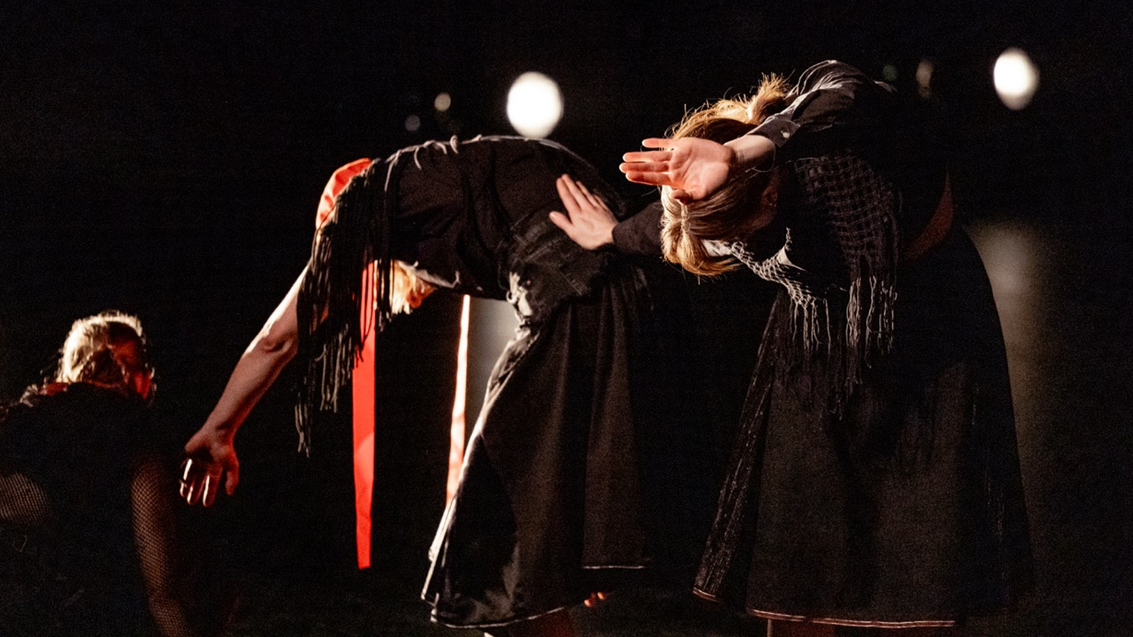 Kuvassa on puoliksi takaa päin taivutusasennossa kaksi tanssijaa mustissa vaatteissa ja heihin osuu valoa. Tausta on tumma. 