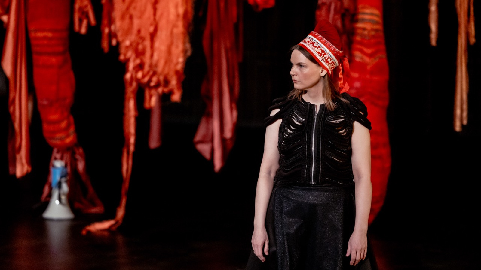 Kuvassa on roikkuvia tumman punaisia tilataideteoksia tummalla taustalla. Oikealla edessä on Elle Sofe Sara seisomassa ja hänellä on musta hihaton mekko ja punainen kirjailtu Lapin päähine päässään. 