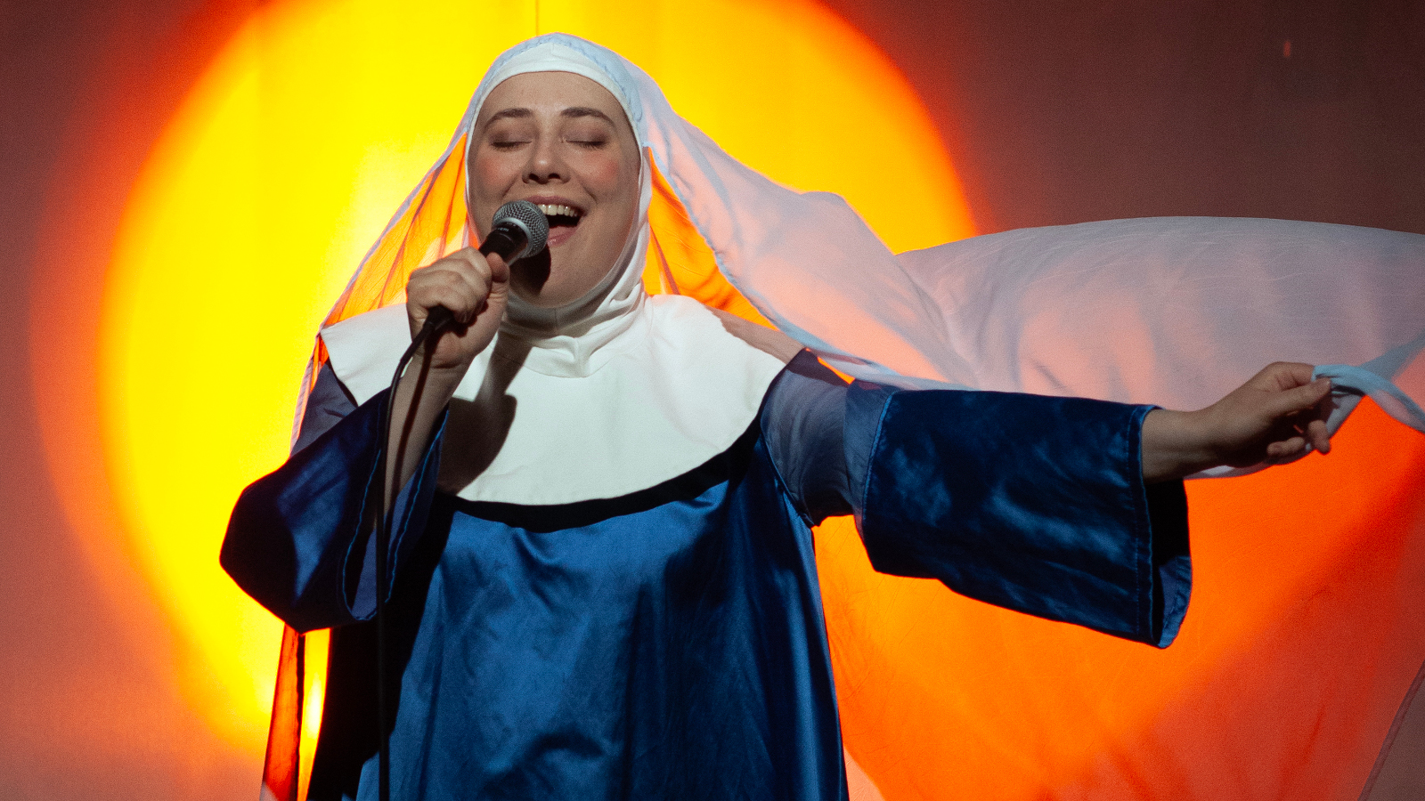 Kuvassa on nainen laulamassa. Hänellä on sininen nunnakaapu, jossa on valkoinen kaulus sekä päähine liehuvine huiveineen. Taustalla on keltainen aurinko ja oranssia valoa.
