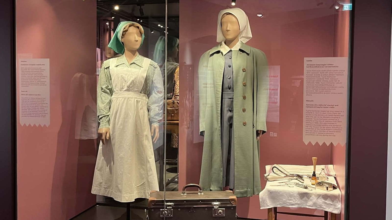 Kuvassa on vitriinissä mallinuket, joiden päällä on vasemmalla on valkoisessa asussa ja mustaraitaisessa puserossa sekä vaalean vihreässä hilkassa. Oikealla on nukella vaalea vihreä takki ja harmaa mekko sekä vaalea hilkka.