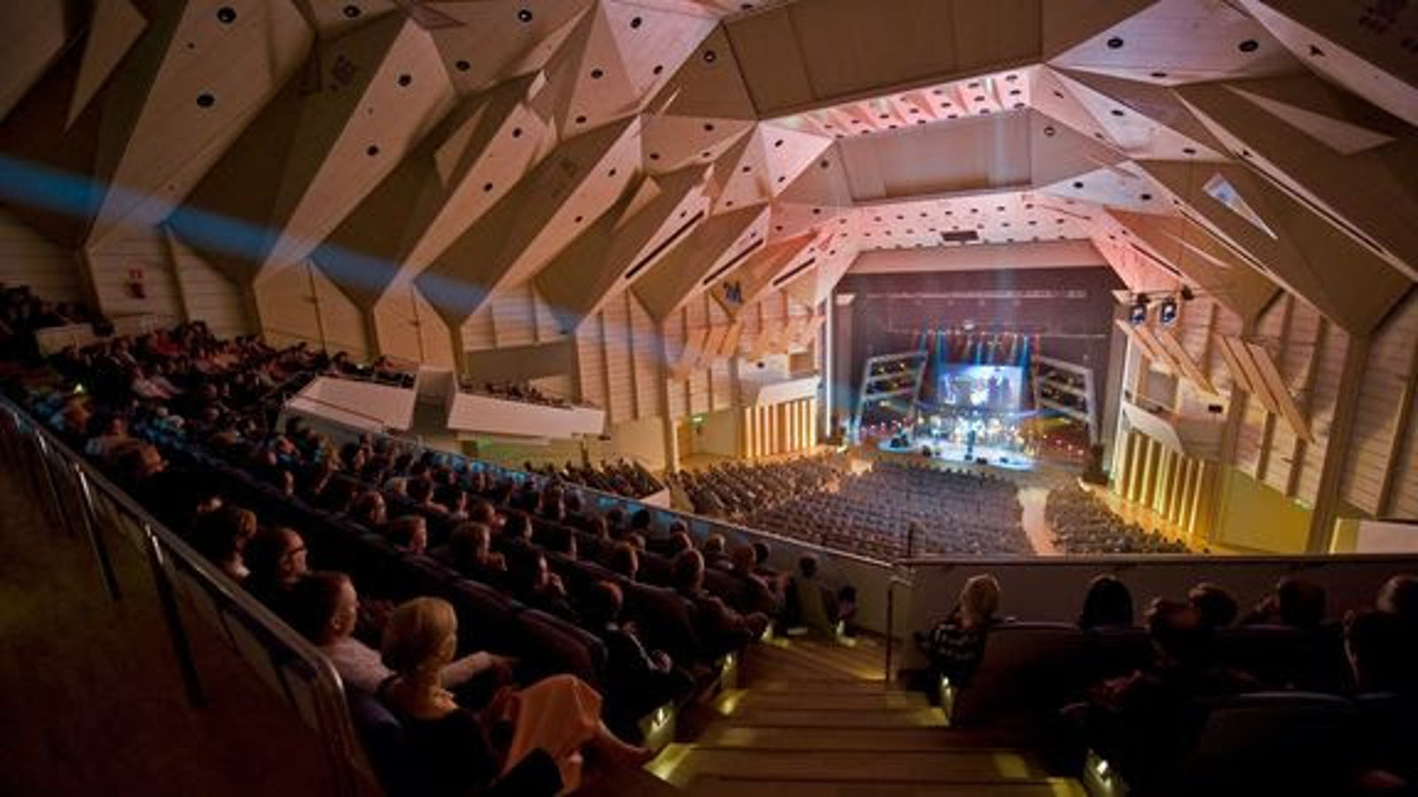 Kuvassa on Tampere-talon konserttisali ylhäältä alaspäin ja hämärässä salissa on yleisöä istumassa  ja katsomassa alhaalla olevalle näyttämölle orkesteria. Vasemmalla ylhäällä on taitettua kattoa.