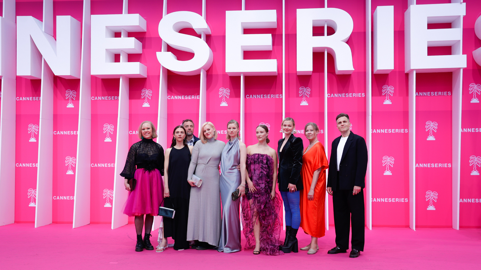 Kuvassa on vaaleanpunaista seinää ja mattoa. Seinällä on isoin valkoisin kohokirjaimin Neseries. Alla ovat suomalaiset yhdeksän näyttelijää eri värisissä juhlapuvuissa.