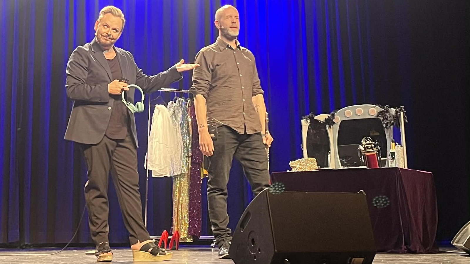 Kuvassa on vasemmalla Mika Tepsa ruskeassa puvussa ja hän esittelee vasemmalla kädelläään Iiro Ollilaa, jolla on myös ruskeat vaatteet päällään. Taustalla on rekki , jossa on naisten vaatteita. Taustalla on sinistä verhoa.