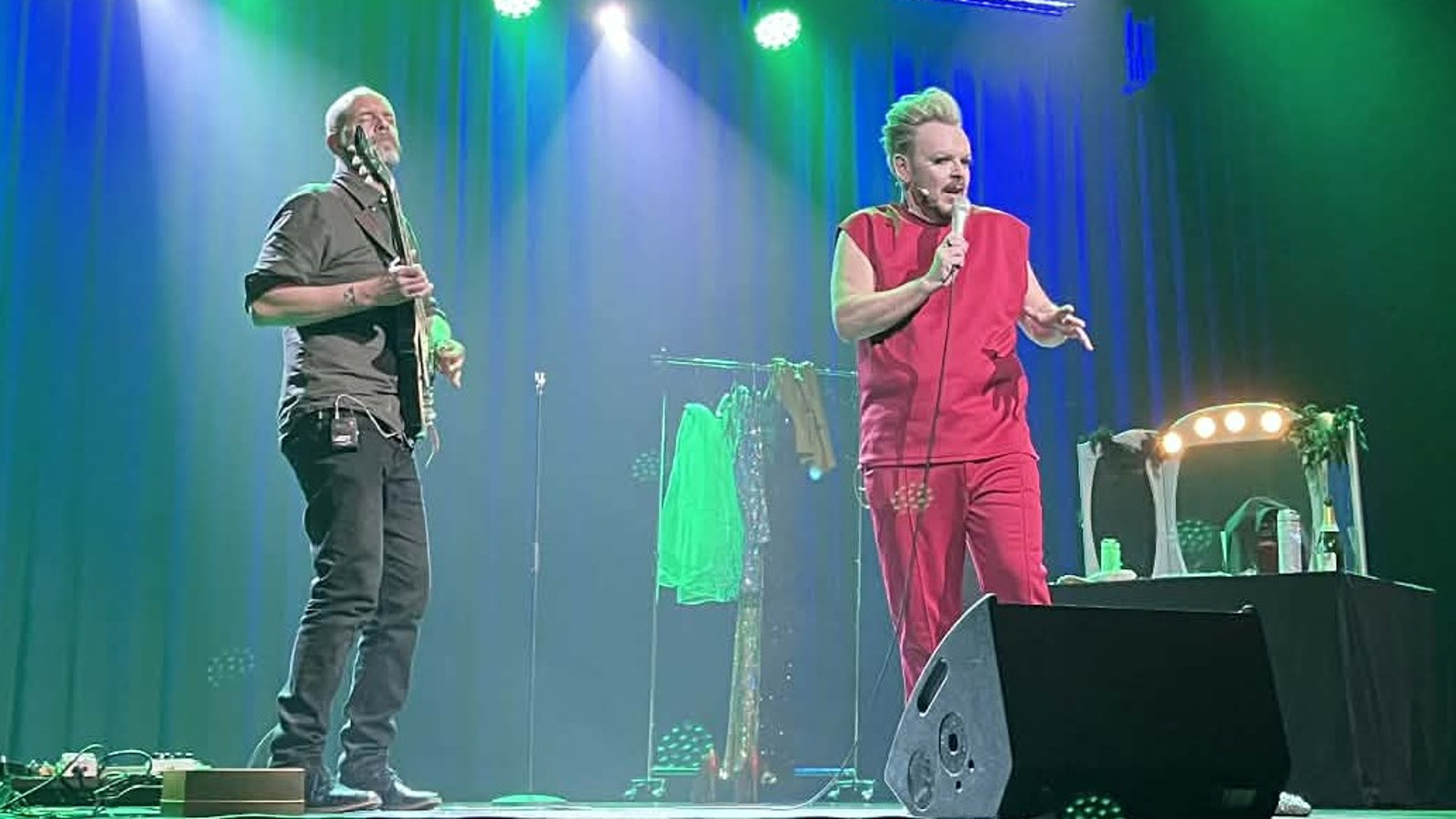 Kuvassa on vasemmalla Iiro Ollila soittamassa kitaraa. Mika Tepsällä oikealla laulamassa punaisessa, hihattomassa puserossa ja pitkissähousuissa.  Taustalla on vaaterekki. Taustalla on sinistä ja vihertävään verhoa.