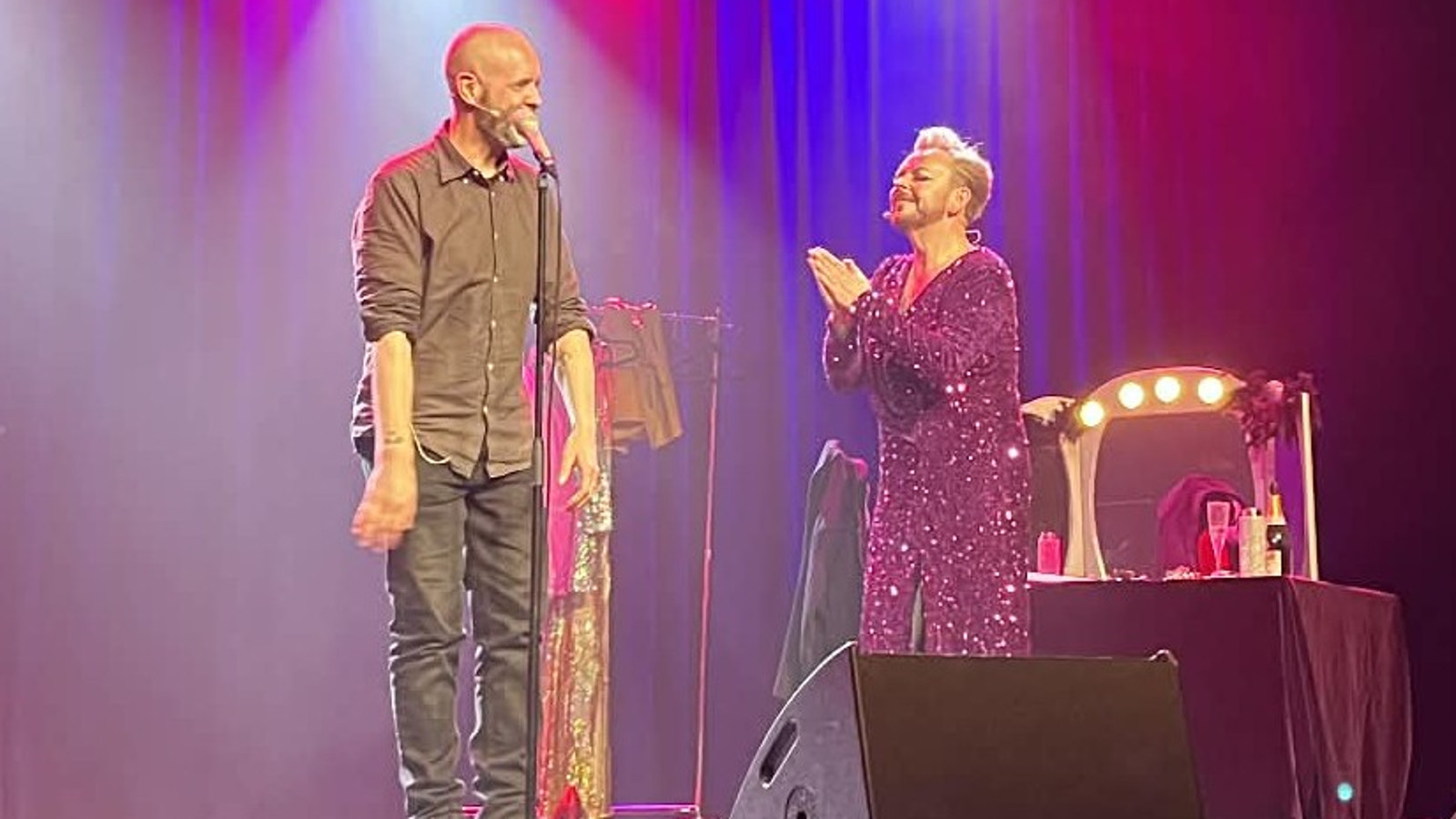 Kuvassa on vasemmalla Iiro Ollila laulamassa mikin ääressä ja oikealla on Mika Tepsä sivuttain ja katsoo kohti Ollilaa kädet yhdessä. Taustalla on sinistä ja punaista valoa.