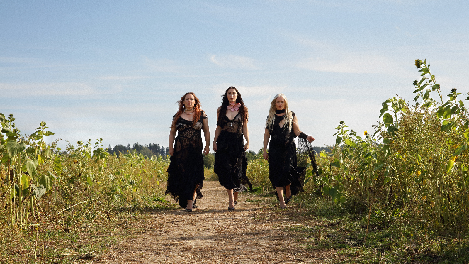 Kuvassa ovat Rodeon kolme jäsentä kävelyllä hiekkatiellä ja vierellä on korkeita kasveja pellolla. Heillä tummat pitkät mekot päällään. Taustalla on sinistä taivasta.