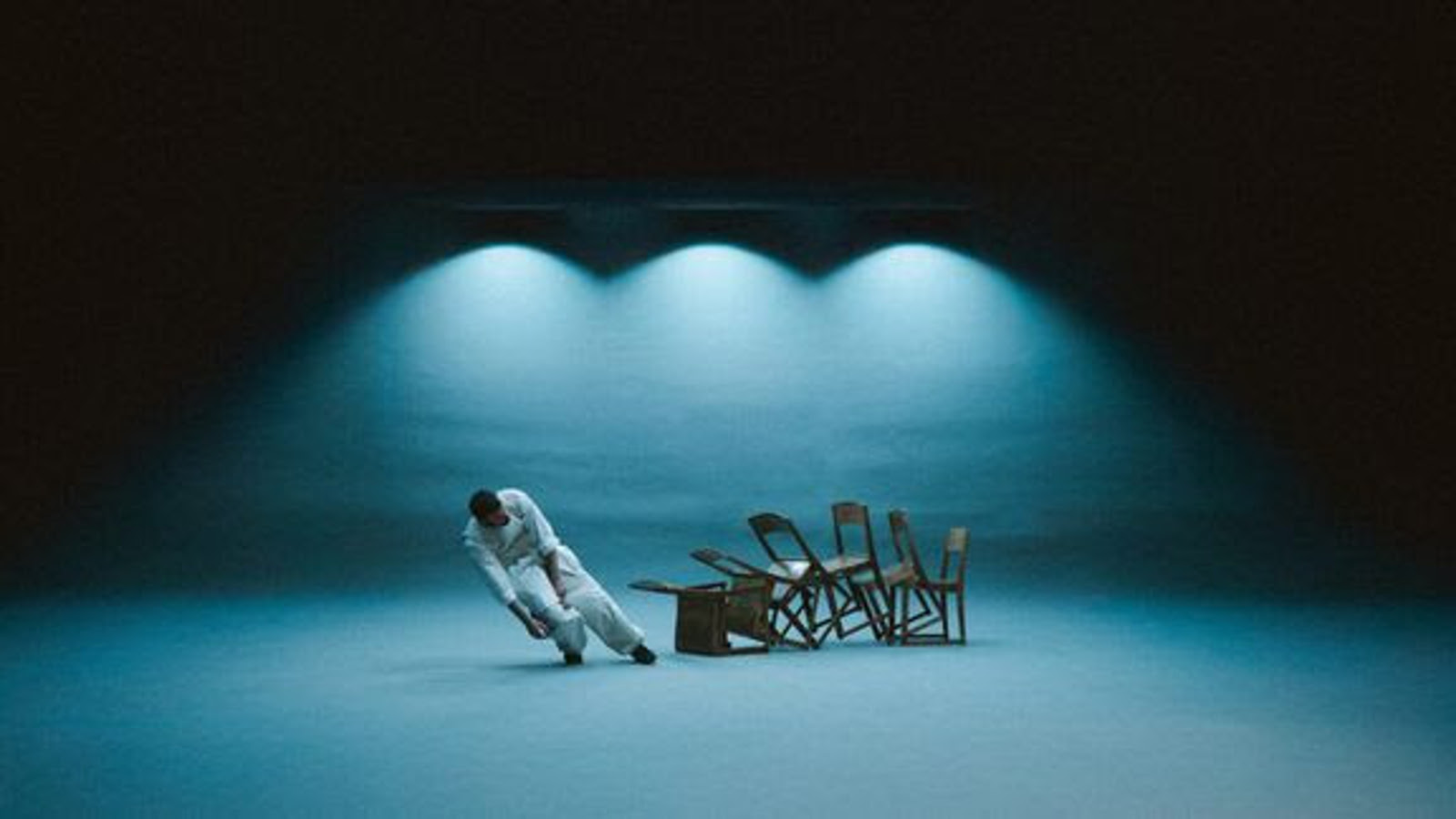 Kuvassa on vasemmalla vinossa asennossa mies valkoisessa haalarissa. Oikealla vieressä on ruskeita puutuoleja vinosti kaatumassa. Tausta on sininen ja yläosastaan musta.