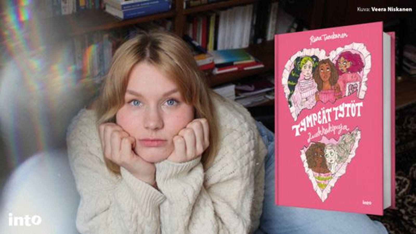 Kuvassa on Riina Tanskanen löhöämässä eteenpäin ja hän nojaa poskiinsa. Hänellä on vaalea villapaita päällään. Oikealla on kuva kirjan kannesta, joka on vaalean punainen. Takana on kirjahyllyä.