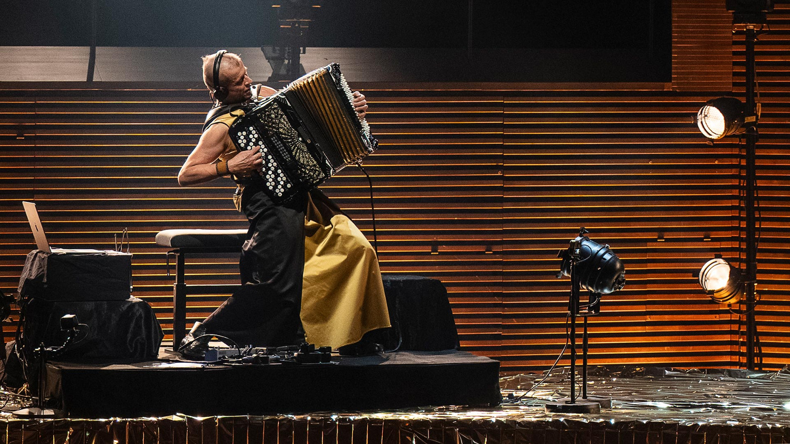 Kuvassa on vasemmalla jalustalla sivuttain Kimmo Pohjonen keltaisissa leveissä housuissa soittamassa harmonikkaa. Takana on ruskeaa vaakasuoraa rimaa.
