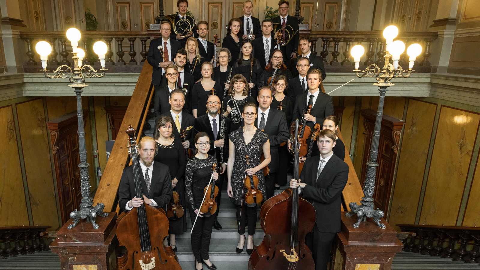 Kuvassa on Vaasan kaupunginorkesterin jäsenet seisomassa leveillä portailla ja taustalla sekä sivuilla on keskiruskeaa panelointi. Ylhäällä on molemmin puolin kolmen lampun valaisimet. 