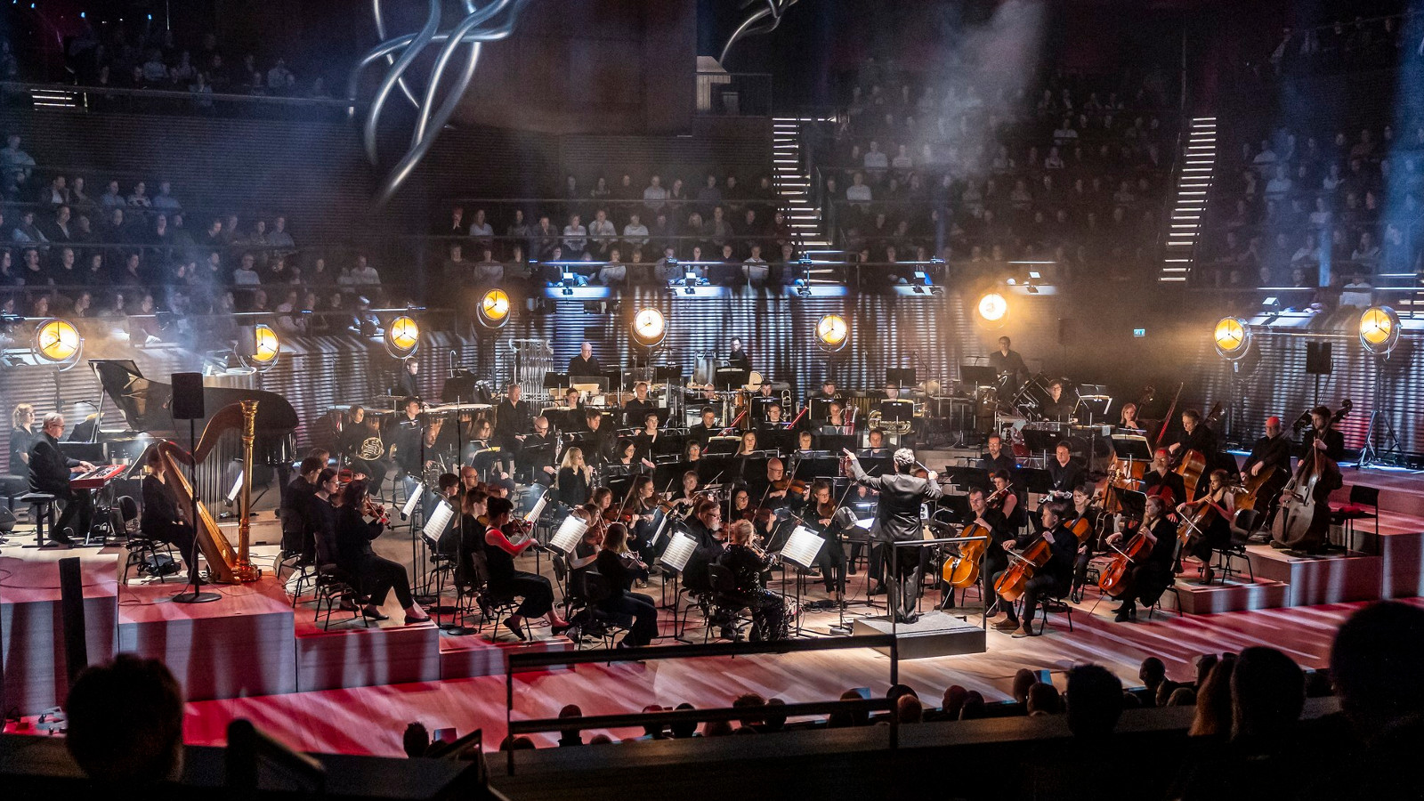 Kuvassa on Vantaan Viihdeorkesteri esiintymislavalla Musiikkitalossa. Taustalla näkyy hämyisessa valaistuksessa yleisöä istumassa nousevassa katsomossa. 