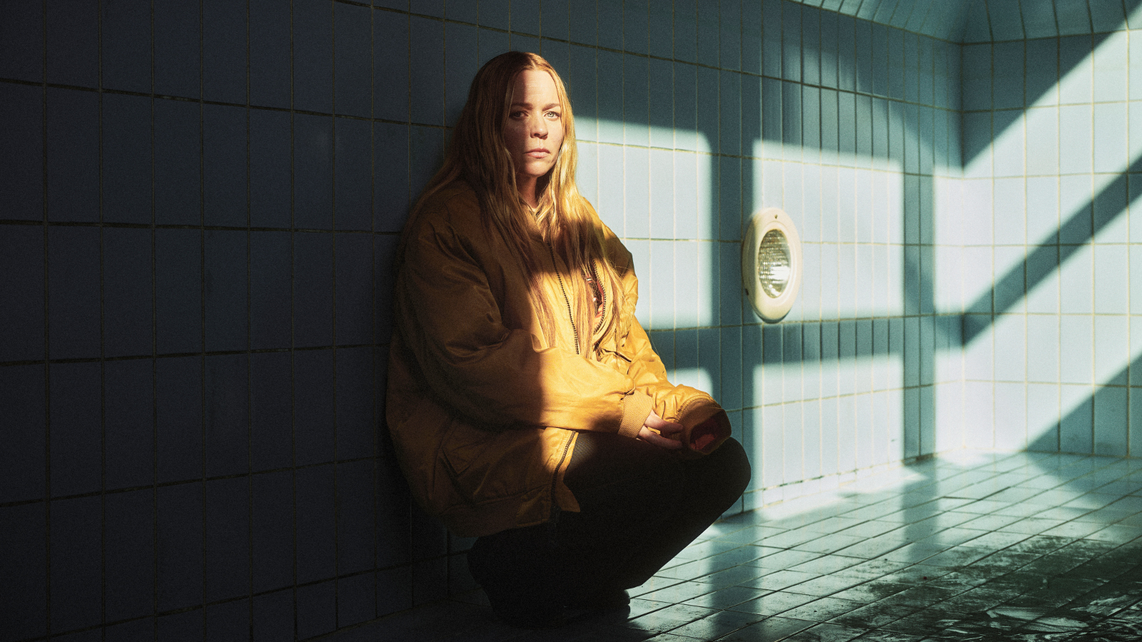   Kuvassa on Paula Vesala kyykussä ja vasen puoli kuvasta on tummaa ja oikealle tulee auringon valoa. Vesala näkyy puolittain. Taustalla on isojen lasi-ikkunoiden nurkka.