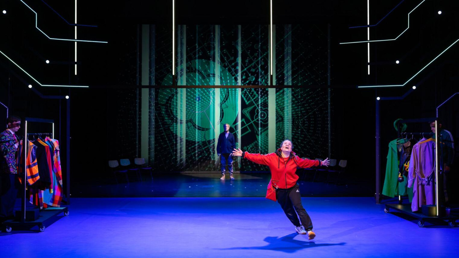 Kuvassa on is tila, jossa sininen lattia ja tummaa taustaa. Takana on vihreän heijastuskuvan edessä on mies. Edessä on vauhdikkaasti tanssiva nainen punaisessa pusakassa.   