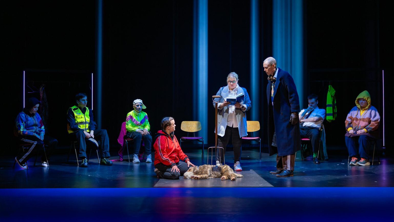Kuvassa on tummalla näyttämöllä edessä mies punaisessa pusakassa polvillaan ja hänen edessään on vaalean ruskea koira. Hänen vieressään seisoo nainen lukemassa kirjasta tekstiä ja hänellä on vaalean sininen jakku. Hänen vierellään oikealla seisoo mies tummassa takissa. Vasemmalla istuu kolme henkilöä ja oikealla kaksi pusakoissa.
