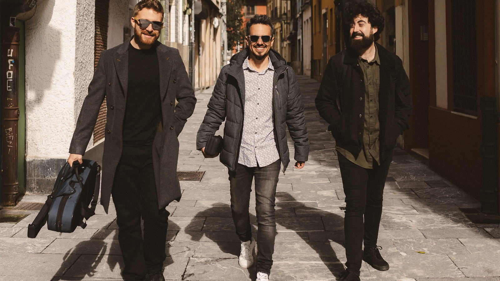 Kuvassa on kolme nuorta miestä kävelemässä kapeaa talojen reunustamaa katua. Heillä n mustat vaatteet paitsi keskimmäisellä harmaa paita.