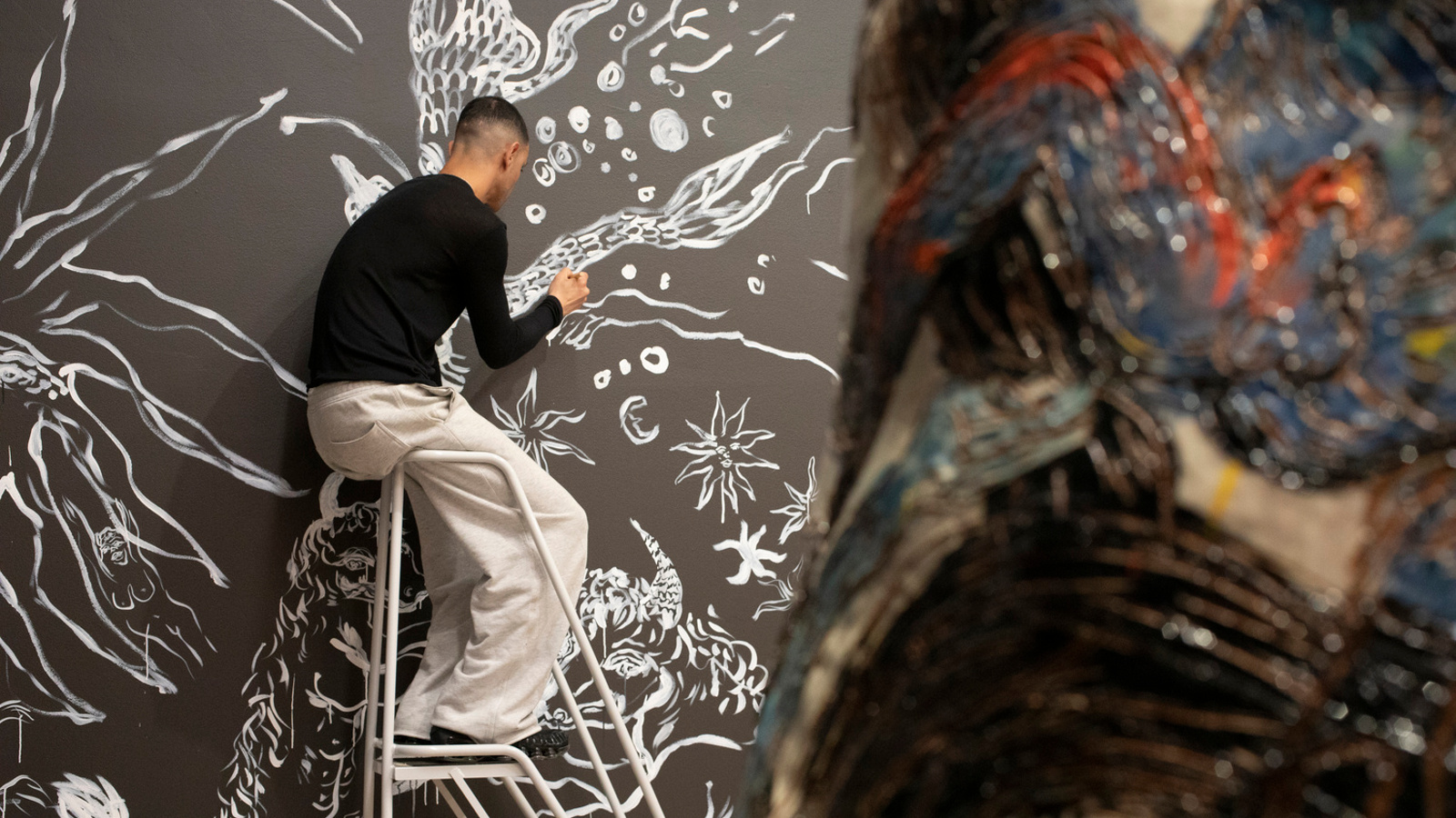 Kuvassa on vasemmalla on korkeiden tikkaiden päällä mustassa puserossa ja valkoisissa housuissa maalamassa ruskealle pohjalle vakoisia isoja kasveja. Oikealla on iso värikäs keramiikkateos.