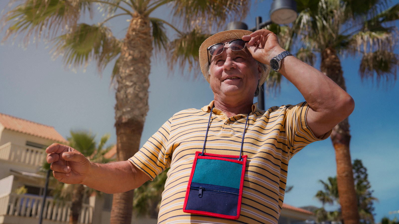 Kuvassa on Tom Lindholm kuvattuna yläviistoon. Hänellä on karttalaukku kaulassaan ja hän pitää aurinkolaseista kiinni vasemmalla kädellään. Taustalla on palmuja, vaaleita rakennuksia ja sinistä taivasta.