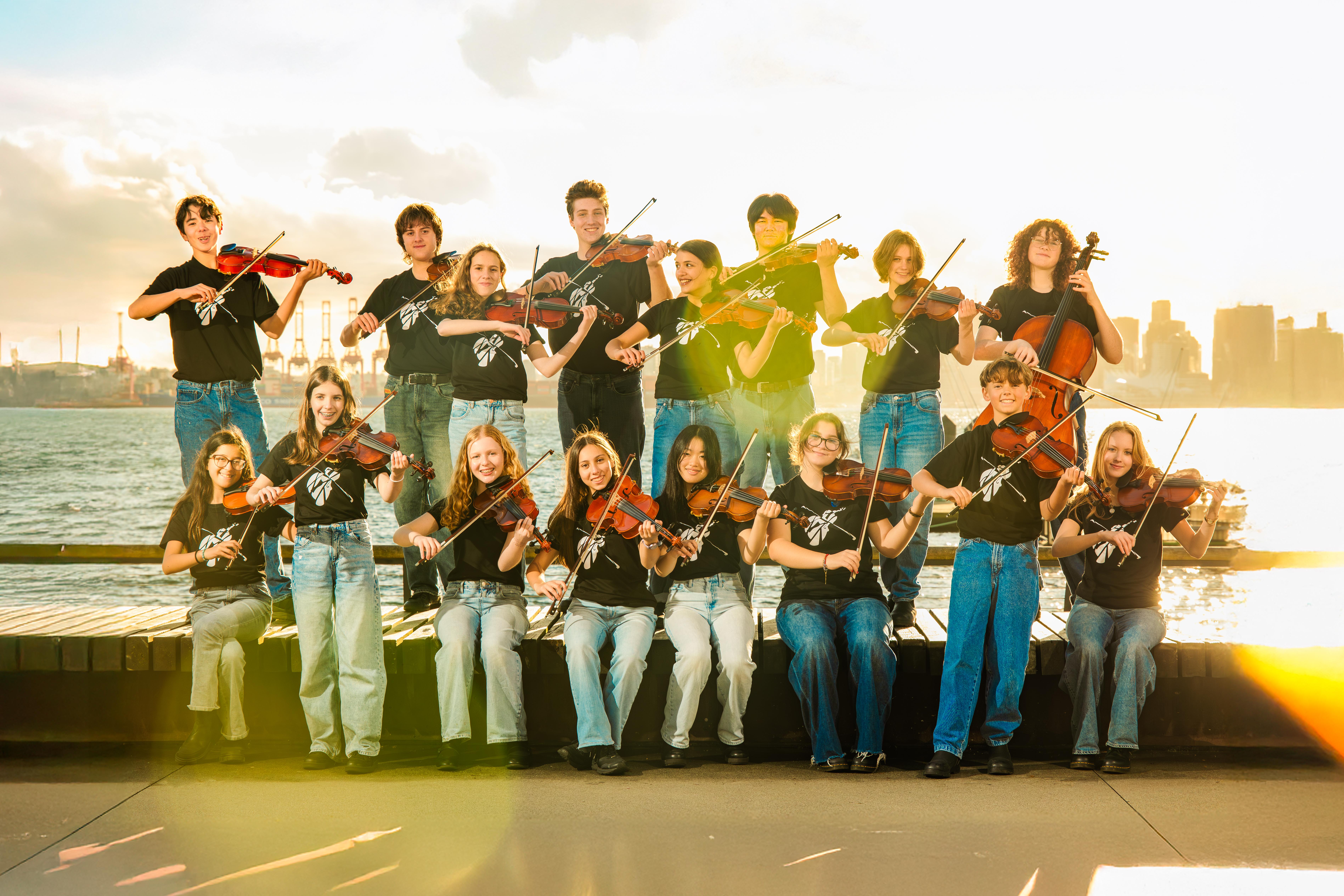 Kuvassa on kahdessa rivissä 20 nuorta soittajaa soittamassa viulua. Taustalla on vesimaisemaa.