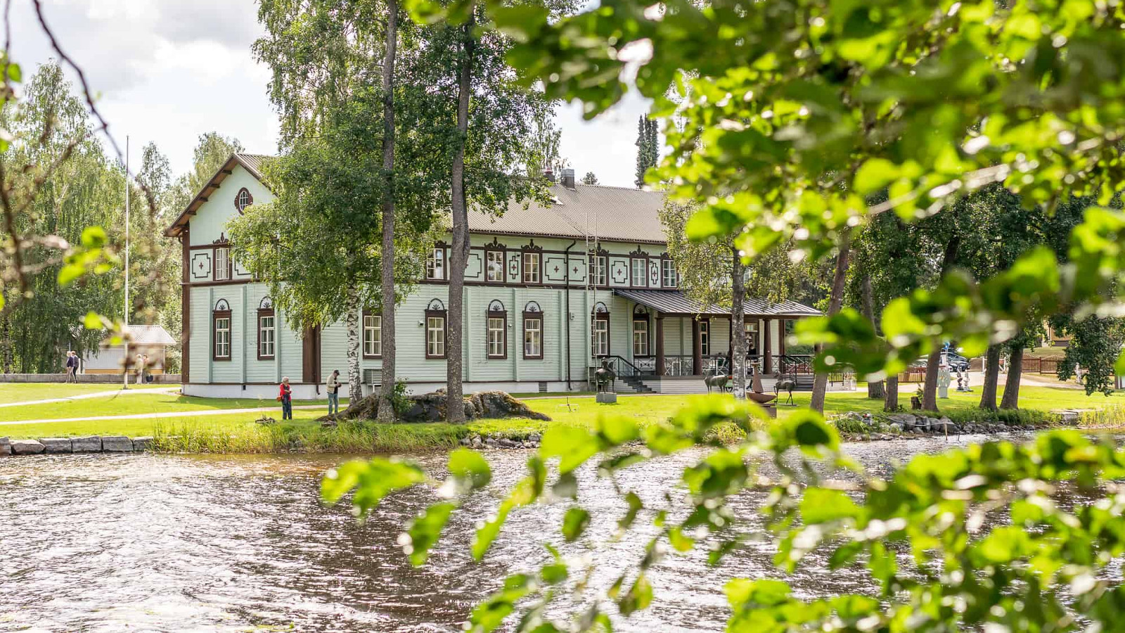 Kuvassa on etualalla järveä ja taaempana on Taidekeskus Salmelan päärakennus, joka on vaalean turkoosi ja siinä on ruskeat ikkunanpielet ja ylhäällä koristeellisia puuleikkauksia. 