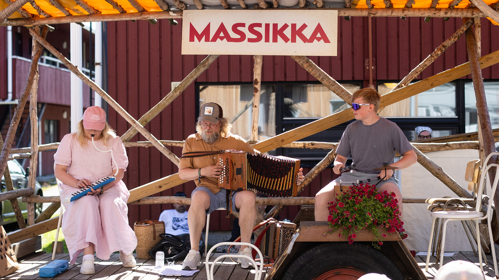 Kuvassa on Vasemmalla nainen vaalean punaisessa mekossa soittamassa melodikaa ja keskellä Antti Paalanen soittamassa harmonikkaa. Vasemmalla on mies soittamassa pieniä rumpupja. He ovat puisen katoksen alla ja taustalla on punainen rakennus.