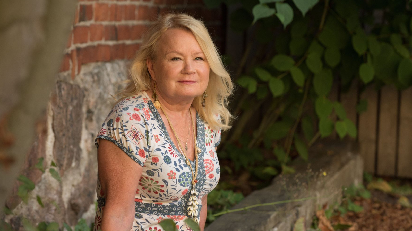 Kuvassa on vasemmalla Kaarina Griffiths istumassa puolivartalokuvassa vaalean kukallisessa mekossa. Taustalla on tiiliseinää ja vihreää pensasta.