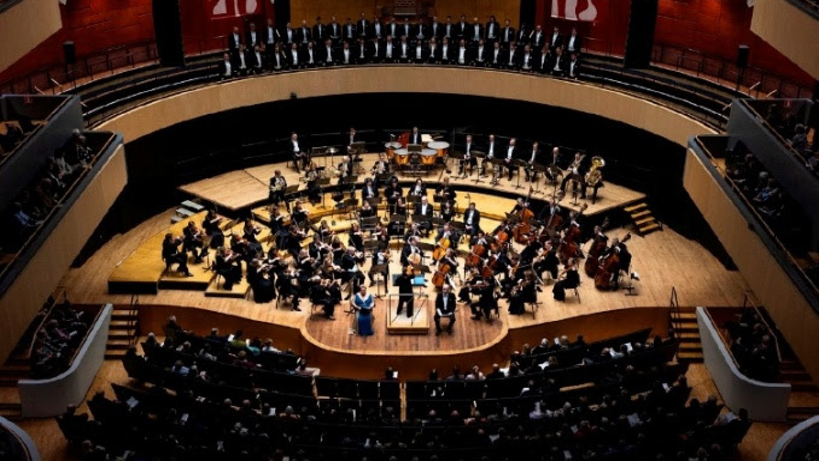 Kuvassa on Sinfonia Lahti ylhäältä kuvattuna soikealla puisella esiintymislavalla soittamassa. Ylempänä näkyy parvekkeella yleisöä ja edessä permannolla. Kuva on osa kirjan kannesta.