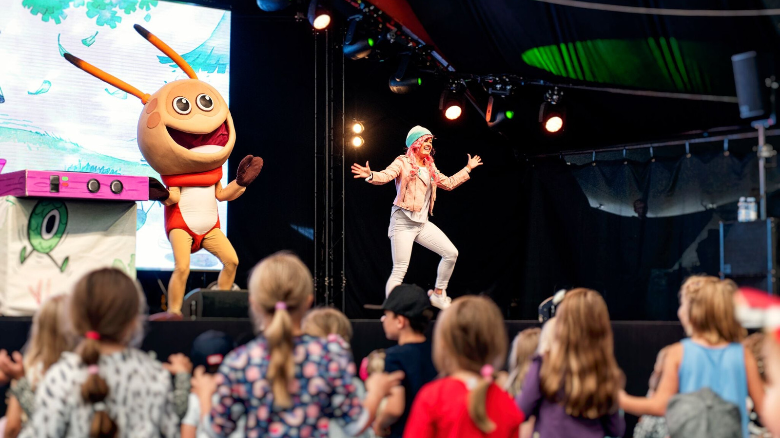 Kuvassa on esiintymislavalla vasemmalla Kikattava Kakkiainen, joka on oranssinen otus isoine päineen ja tuntosarvineen. Silla on punainen paita päällään. Lavalla oikealla on naislaulaja, jolla vaaleat vaatteet ja hän tekee loikkaa kädet levitettyinä.