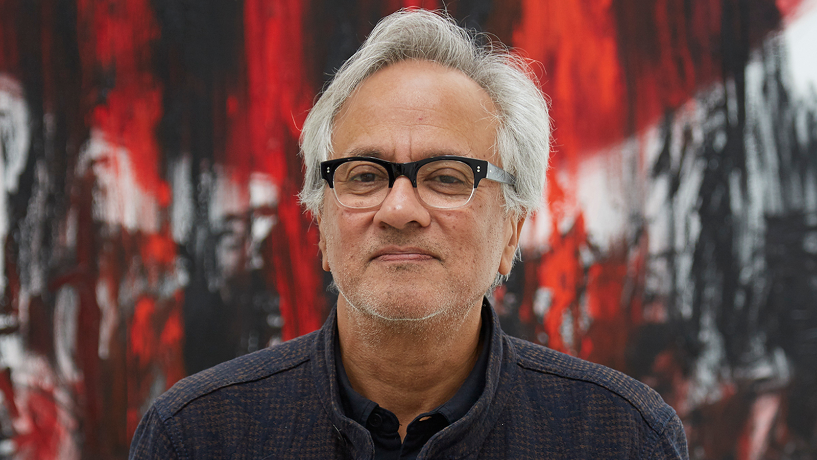 Kuvassa on Anish Kapoor puolivartalokuvassa ja hänellä on vaaleat hiukset sekä tumma paita. Taustalla on mustan punaista teosta.