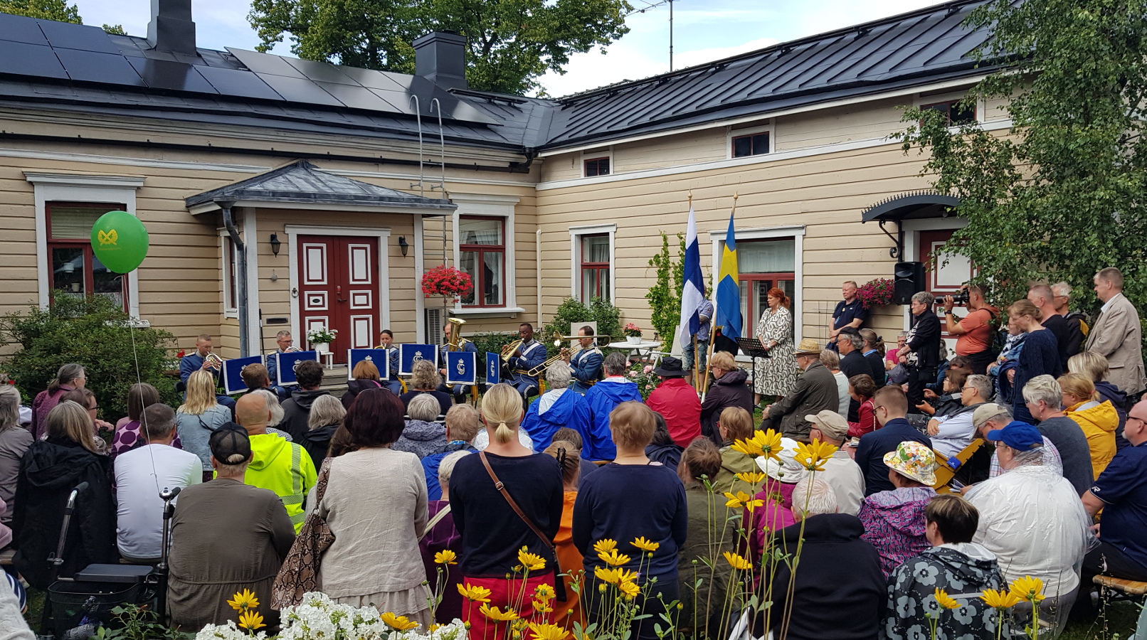 Kuvassa on etualalla yleisöä kuuntelemassa penkeissä istuen ja takana on kellertävä puutalo, joka tekee kulman. Edessä soittokunta soittamassa.