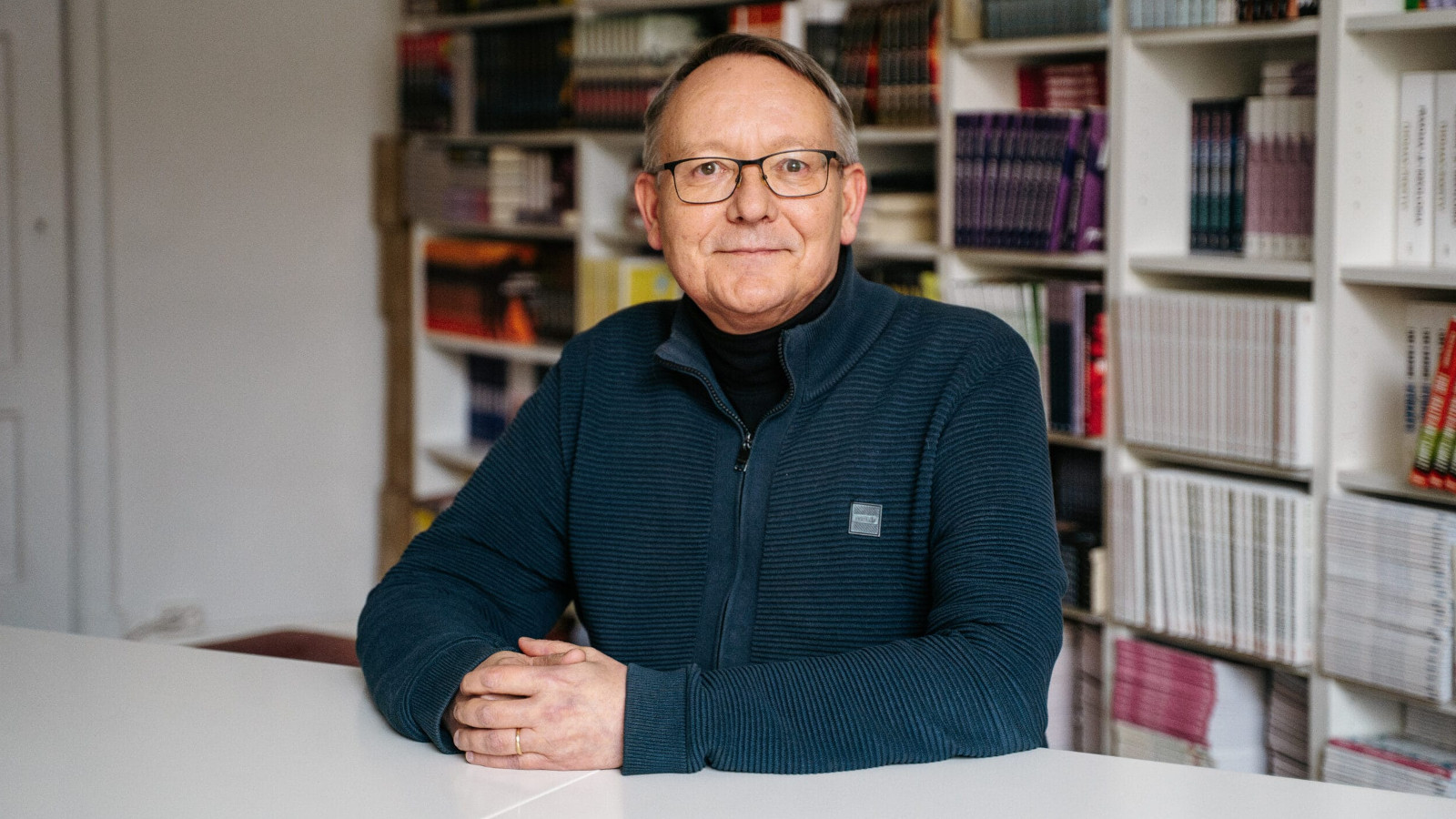 Kuvassa on Juha-Antero Puistola puolivartalokuvassa istumassa vaalean pöydän ääressä. Hänellä on musta pusero päällään. Taustalla on kirjahyllyä.